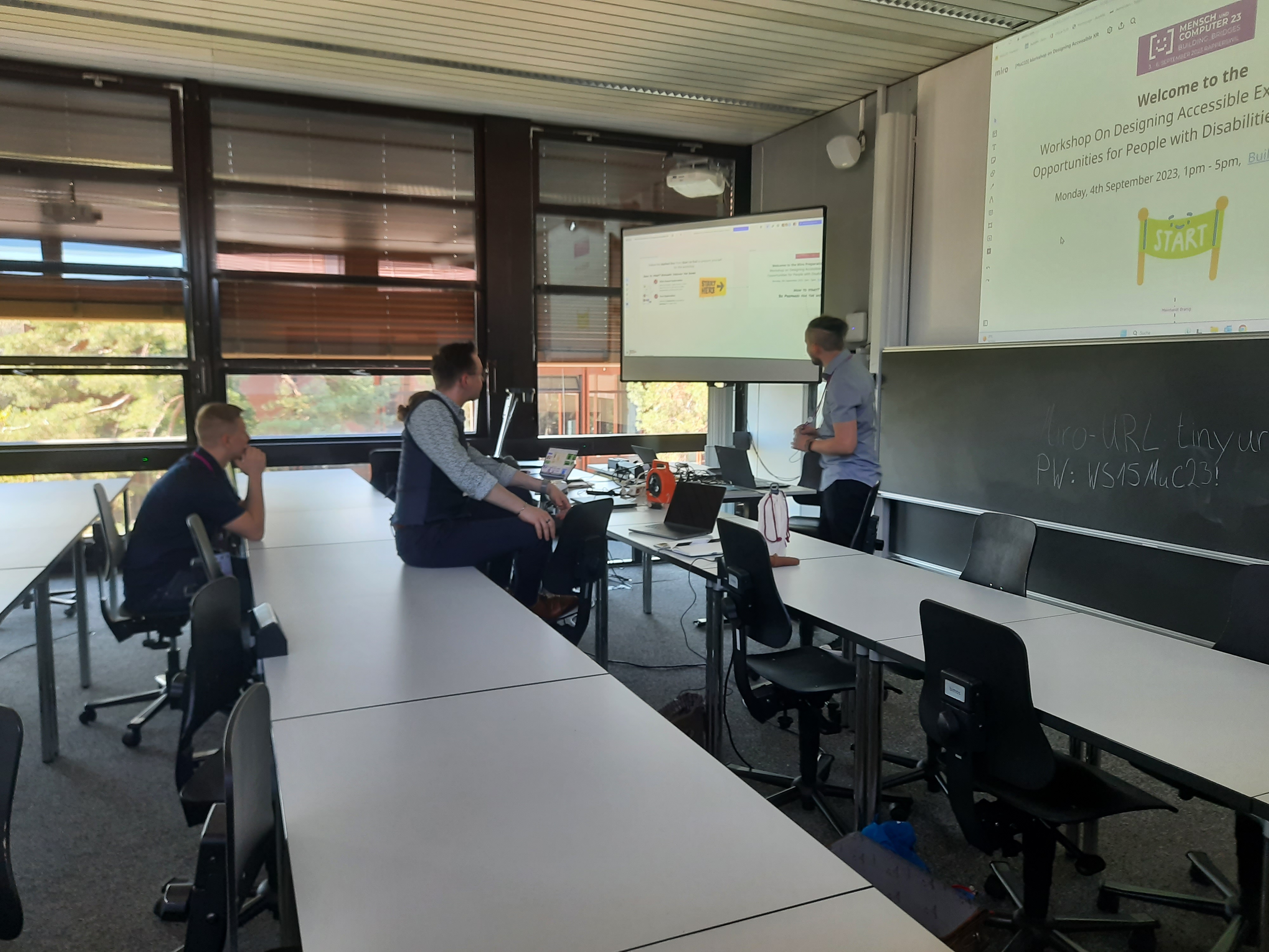 2 Personen sitzen in der ersten Reihe, eine steht vor Beamer mit Folien auf der steht: 