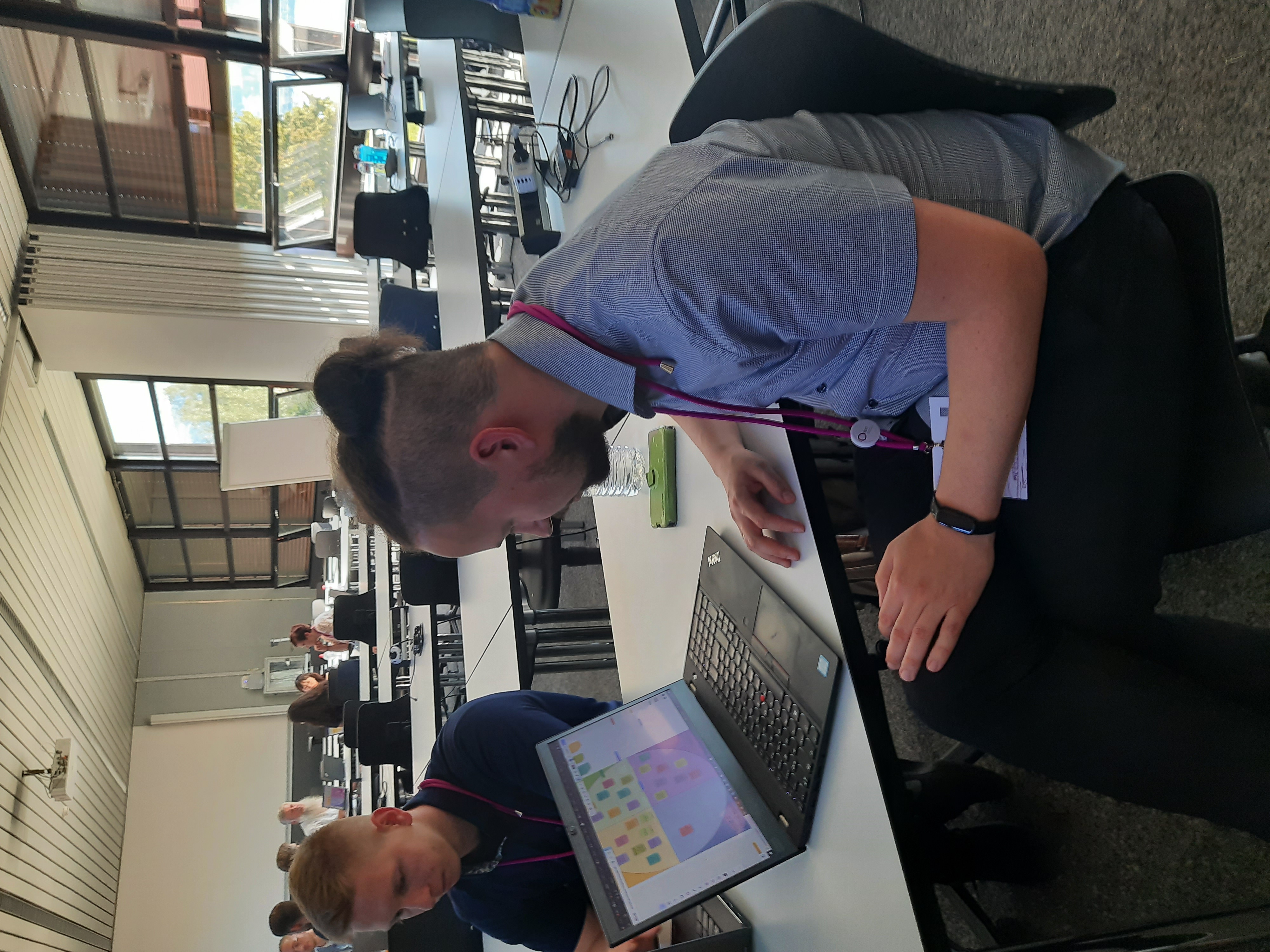 Zwei Personen sitzen an einem Tisch, eine davon schaut in den Laptop vor ihm. Im Hintergrund sitzen weitere Personen im Kreis und diskutieren.