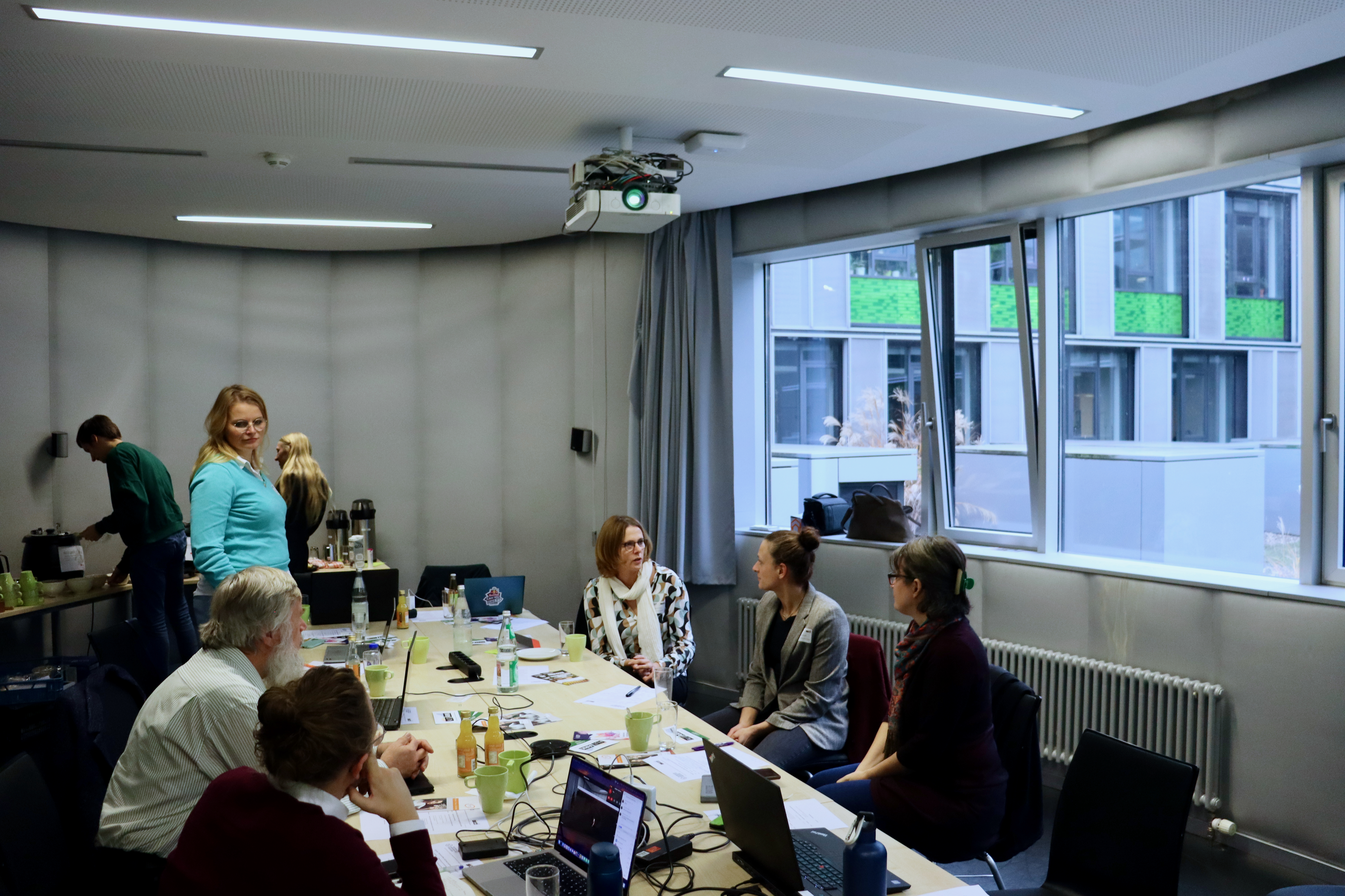 Foto des Veranstaltungsraumes in lockerer Atmosphäre: Eine weiblich gelesene Person steht, rechts davon sitzen zwei weiblich gelesene Personen, die sich ausgiebig unterhalten, mehrere Personen hören zu. Im Hintergrund ist ein Buffet aufgebaut, 2 Personen stehen davor.