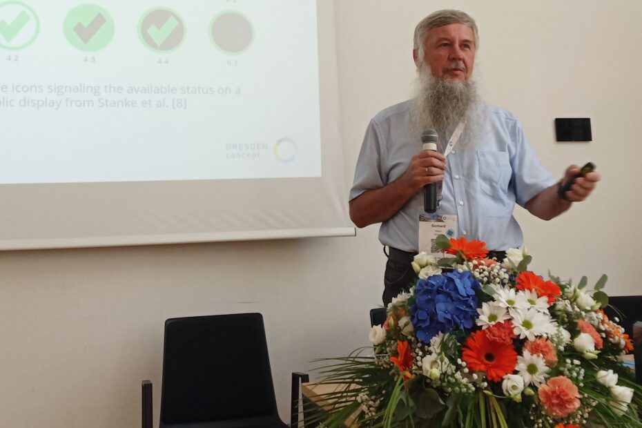 Prof. Weber stands in front of a set of slides projected onto the wall with a microphone in his hand and speaks. The slides show various symbols for barriers in the workplace.
